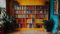 Colorful bookshelf with rainbow-organized books against vibrant yellow wall, flanked by potted plants on wooden floor in cozy reading nook.