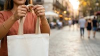 Woman in coral shirt holding white canvas tote bag on urban street with blurred pedestrians and buildings in background, suitable for retail, shopping, and eco-friendly lifestyle marketing.
