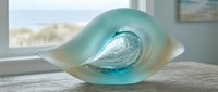 Translucent turquoise glass wave sculpture on wooden surface near window with ocean view. Modern coastal home decor accent piece for contemporary interior design.