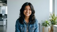 Young woman with long dark wavy hair and warm smile wearing denim shirt in modern office environment with plants, suitable for corporate, business, and professional lifestyle content.