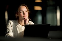 Young Caucasian woman in white sweater working on laptop at night, illuminated by warm light, with thoughtful expression resting chin on hand.