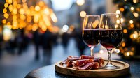 Two glasses of red wine with charcuterie on wooden board against warm bokeh lights creating romantic evening atmosphere for dining and celebration.