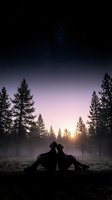 Silhouetted couple sitting back-to-back in misty forest clearing at sunrise with coniferous trees and starry sky, conveying intimacy and connection in nature for romantic and lifestyle content.