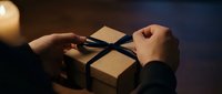 Hands unwrapping kraft paper gift box tied with black ribbon against dark background with warm lighting.