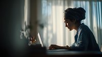 Young African American woman with dreadlocks working on laptop computer in modern office space with natural window lighting for business and technology concepts.