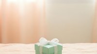 Green gift box with white ribbon bow on wooden surface against blurred beige background, suitable for holiday, celebration, and promotional design projects.