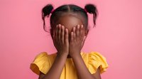 Young African American girl in yellow dress covering face with hands against pink background, expressing shyness, playfulness or hide and seek emotions.