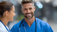 Smiling male doctor in blue scrubs with stethoscope talking to female colleague in medical facility. Healthcare professionals discussing patient care in hospital setting.