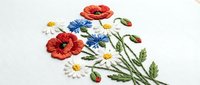 Delicate hand embroidered wildflower bouquet with red poppies, white daisies and blue cornflowers on white fabric. Traditional needlework craft for home decor.