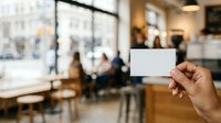 Hand holding blank white card in modern office lobby with blurred colleagues and city view in background, suitable for business branding and corporate identity mockups.