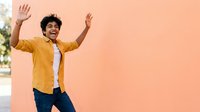 Young man with dark curly hair and medium brown skin tone wearing yellow shirt and jeans, arms raised in celebration against peachy wall background.