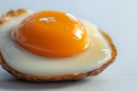 Macro shot of sunny side up fried egg with bright orange yolk and crispy brown edges on white surface, showcasing appetizing breakfast food preparation.