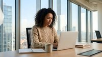 Young woman with curly dark hair and medium skin tone working at wooden desk with laptop, coffee mug, and tablet in modern office with city skyline views through floor-to-ceiling windows.
