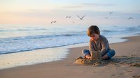 Young boy playing with sand on peaceful ocean beach at sunset with seagulls flying overhead. Child enjoying summer vacation by the sea during golden hour.