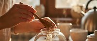 Woman's hands adding chocolate chips to glass jar in warm sunlit kitchen for homemade baking preparation and cooking ingredients storage.