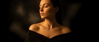 Elegant young woman with closed eyes in off-shoulder black dress against dark background with warm golden lighting for luxury fashion and beauty concepts.