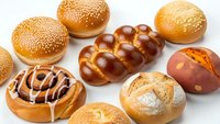 Assorted fresh baked bread rolls and pastries with sesame seeds, braided challah, and glazed sweet buns arranged on white background for bakery display.