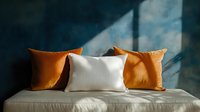 Minimalist home decor with orange and white throw pillows on beige sofa against moody blue wall with natural window light and shadow play.