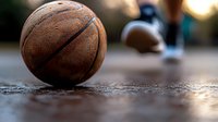 Worn leather basketball resting on asphalt court surface with blurred player in background during golden hour lighting for sports and recreation themes.