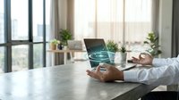 Businessman holding laptop with digital hologram globe display, modern office workspace with natural light and plants, technology and global business concept.