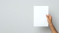 Adult male hand with medium skin tone holding blank white paper sheet against neutral gray wall background, suitable for mockup, advertising, and presentation purposes.