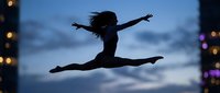 Young woman dancer silhouette performing split leap against twilight sky with city lights, expressing freedom and athletic grace in urban evening setting.