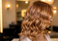 Professional hairstylist showcasing golden brown balayage waves in modern salon with warm lighting for hair color and styling services.