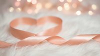 Soft pink satin ribbon forming heart shape on white wooden surface with warm bokeh lights creating romantic Valentine's Day atmosphere for love celebration.