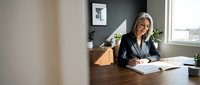 Middle-aged woman with light brown hair, Caucasian ethnicity, wearing professional black blazer and glasses, seated at wooden desk in modern office environment, writing in notebook with plants and cityscape visible in background.