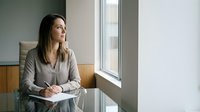 Professional businesswoman sitting at modern office desk looking thoughtfully out window during daytime work session.