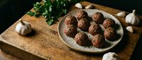 Homemade meatballs on rustic plate with fresh herbs and garlic on wooden cutting board for cooking and recipe content.