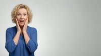Middle-aged woman with curly blonde hair wearing blue shirt, hands on face expressing surprise and excitement against neutral background.