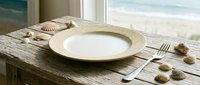 Coastal dining setup with empty plate, seashells, and ocean view through window on rustic wooden table for restaurant, hospitality, and seaside dining concepts.