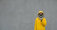 Person in bright yellow hoodie and cap speaking through megaphone against concrete wall background for marketing and communication concepts.