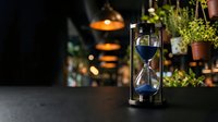 Vintage hourglass with blue sand on dark surface in cozy cafe interior with warm lighting and green plants creating atmospheric business concept.