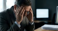 Stressed businessman holding head in hands at office desk with computer screens showing work pressure and mental fatigue in corporate environment.