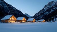 Illuminated wooden chalets in snowy mountain valley at dusk, cozy winter lodges with warm glowing windows against dramatic alpine peaks and pristine snow landscape.