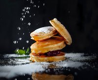 Stack of fresh pancakes with jam filling and powdered sugar falling on dark background with reflection. Food photography with dramatic lighting.