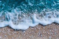 Aerial view of turquoise ocean waves crashing on sandy beach with white foam, creating dynamic pattern of water meeting shoreline. Natural summer seascape background.