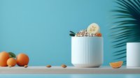 Creamy smoothie bowl topped with banana slices, nuts and chocolate in white ceramic cup on wooden surface against turquoise wall with palm shadow.