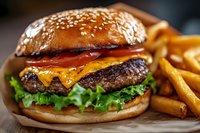 Juicy cheeseburger with melted cheddar, fresh lettuce, tomato on sesame seed bun served with golden french fries, close up view on wooden surface.