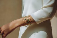 Elegant gold bracelets with emerald gemstone adorning woman's wrist against white blouse, showcasing minimalist luxury jewelry styling.