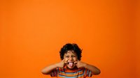 Young boy with dark curly hair and medium skin tone wearing striped shirt, smiling with hands near face against vibrant orange background for portrait and advertising use.