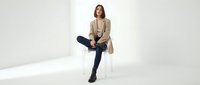 Young professional woman in beige blazer and dark jeans sitting on white chair against neutral background for business and lifestyle concepts.