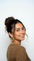 Young Hispanic woman with messy bun hairstyle and gold earrings smiling at camera in brown knit sweater isolated on white background for lifestyle content.