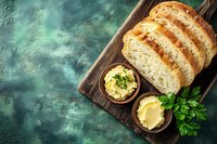 Fresh sliced artisan sourdough bread on rustic wooden board with herb butter in wooden bowls, garnished with parsley against turquoise background.