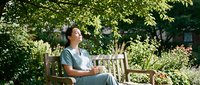 Young Asian woman in light blue dress relaxing on wooden garden bench under green tree canopy, peaceful outdoor meditation moment in sunny backyard setting.