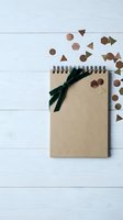Kraft paper notebook with dark green ribbon and geometric confetti on white wooden background for creative projects and stationery design.