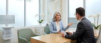 Professional blonde businesswoman consulting with male client in modern bright office space with large windows and contemporary furniture.