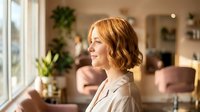 Young blonde Caucasian businesswoman in beige blazer smiling while looking out window in modern office space with plants and natural lighting for corporate content.